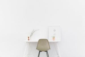 A minimalist home office with white wall, white desk, chair, small vase, white picture.